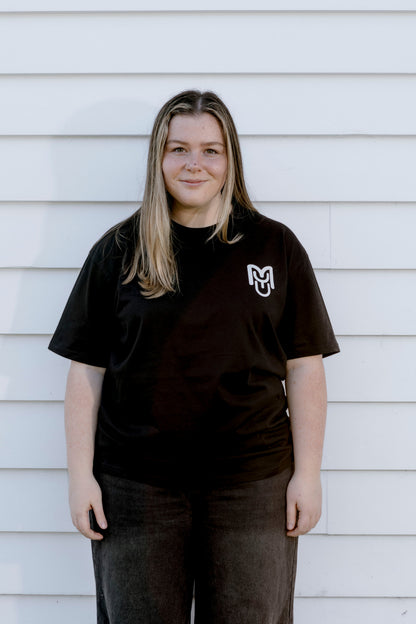 A woman with blonde-highlighted hair stands against a white horizontal siding wall, wearing a black short-sleeve t-shirt with a white stylized 'M' logo on the left chest and dark gray pants.