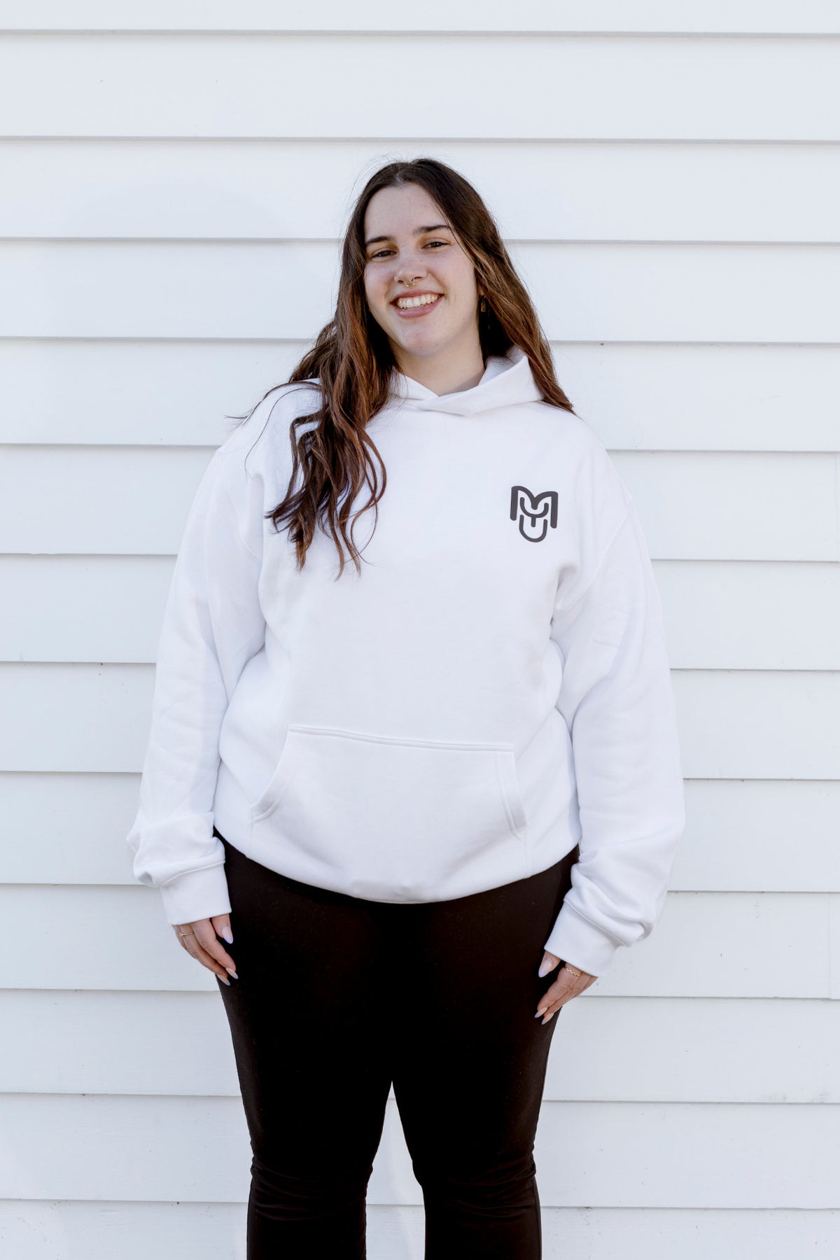 A smiling young woman with long brown hair stands against a white horizontal siding wall, wearing a white pullover hoodie with a small black stylized 'M' logo on the left chest and dark brown pants.